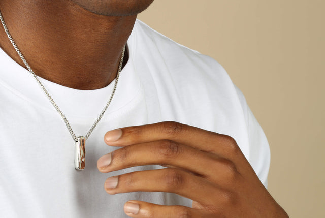 a man wearing a white T shirt and wearing a liquid block necklace in silver. His hand is reaching to touch it.